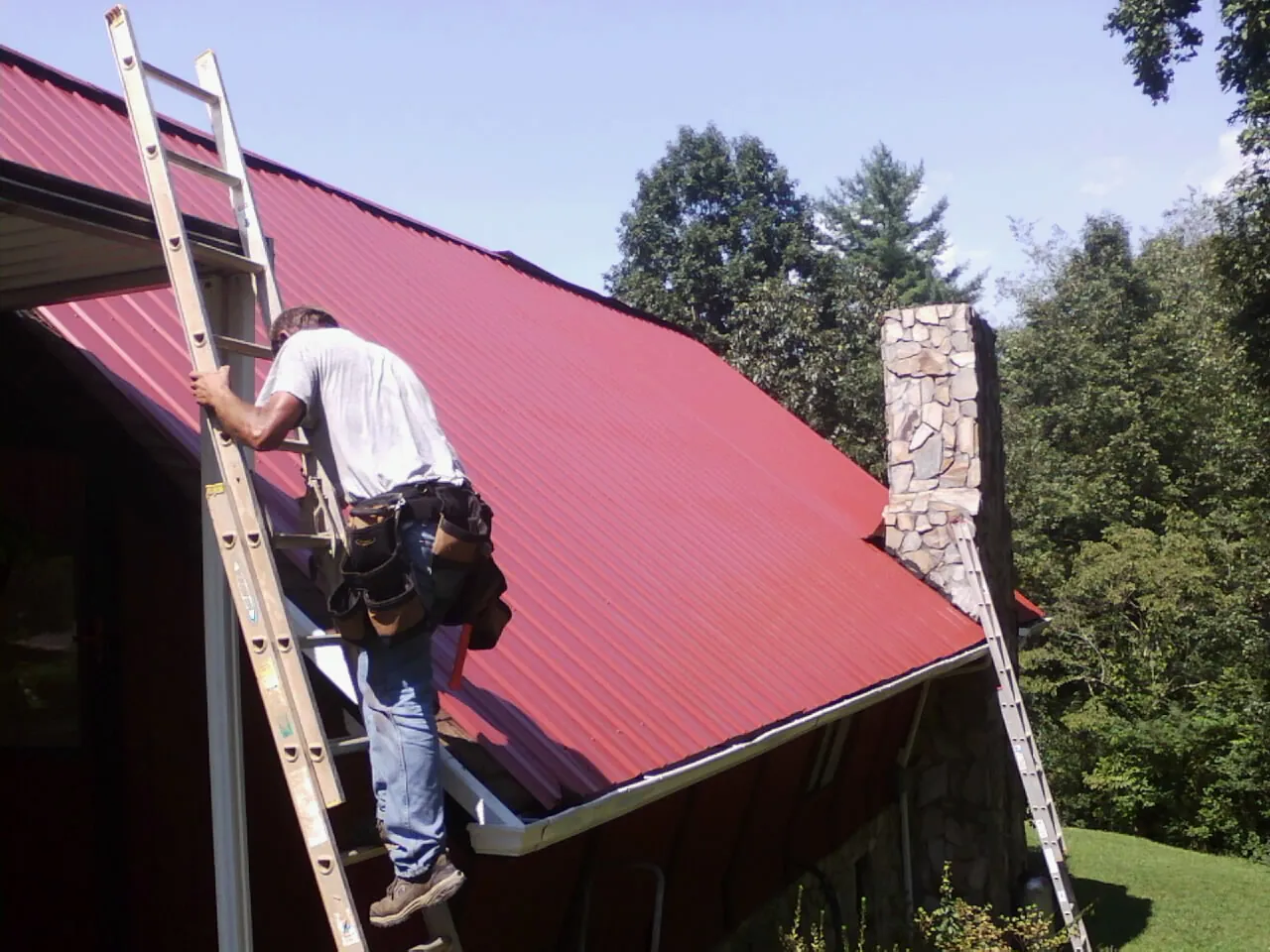 Roofer on a ladder beside a red metal roof and stone chimney — Soffit & Fascia Repair by Roofing Brothers in Peterboro